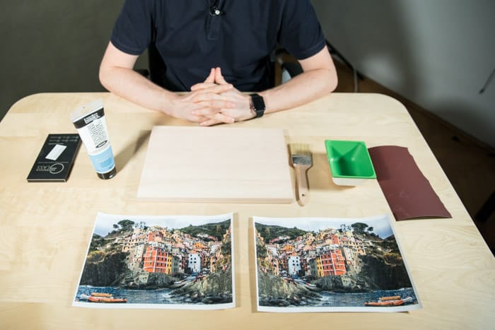 一名男子坐在一张木桌，在他是两张明亮的沿海城镇，胶水，画笔，砂纸等材料的两个相同的照片，将激光印刷到木材
